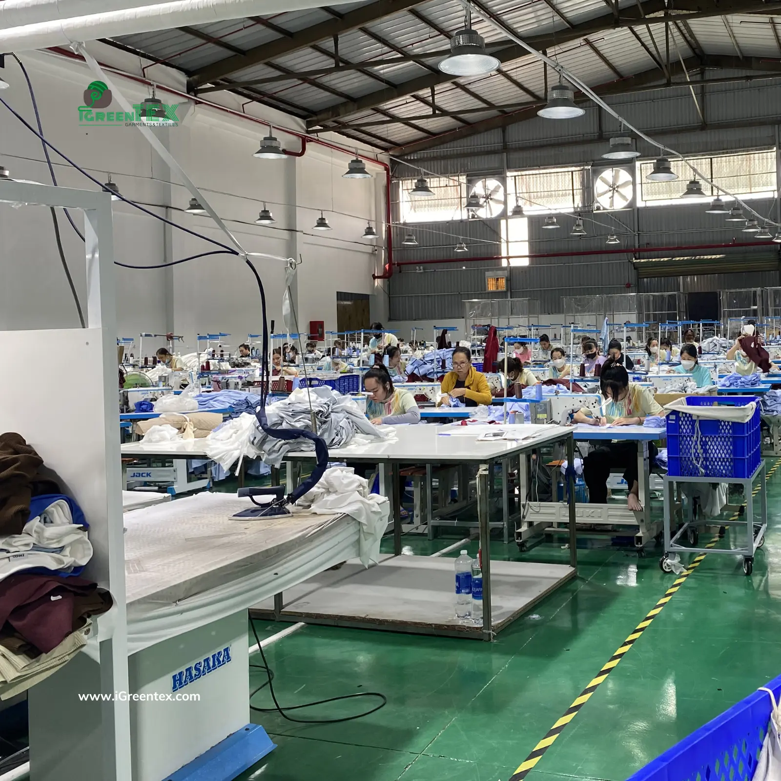 A wide-angle photograph capturing the dynamic and organized sewing floor of iGreen Tex Vietnam, a premier garment manufacturer in Vietnam. Hundreds of dedicated employees, wearing standardized uniforms (referencing previous image image_25.png and image_32.png), are focused and working hard on multiple active sewing lines. Hanging factory signage reinforces technical compliance, providing unedited visual proof of scale and operational dedication directly satisfying the need to verify garment manufacturer in vietnam for international export. The perspective captures the vast, active production environment directly in Ho Chi Minh City.