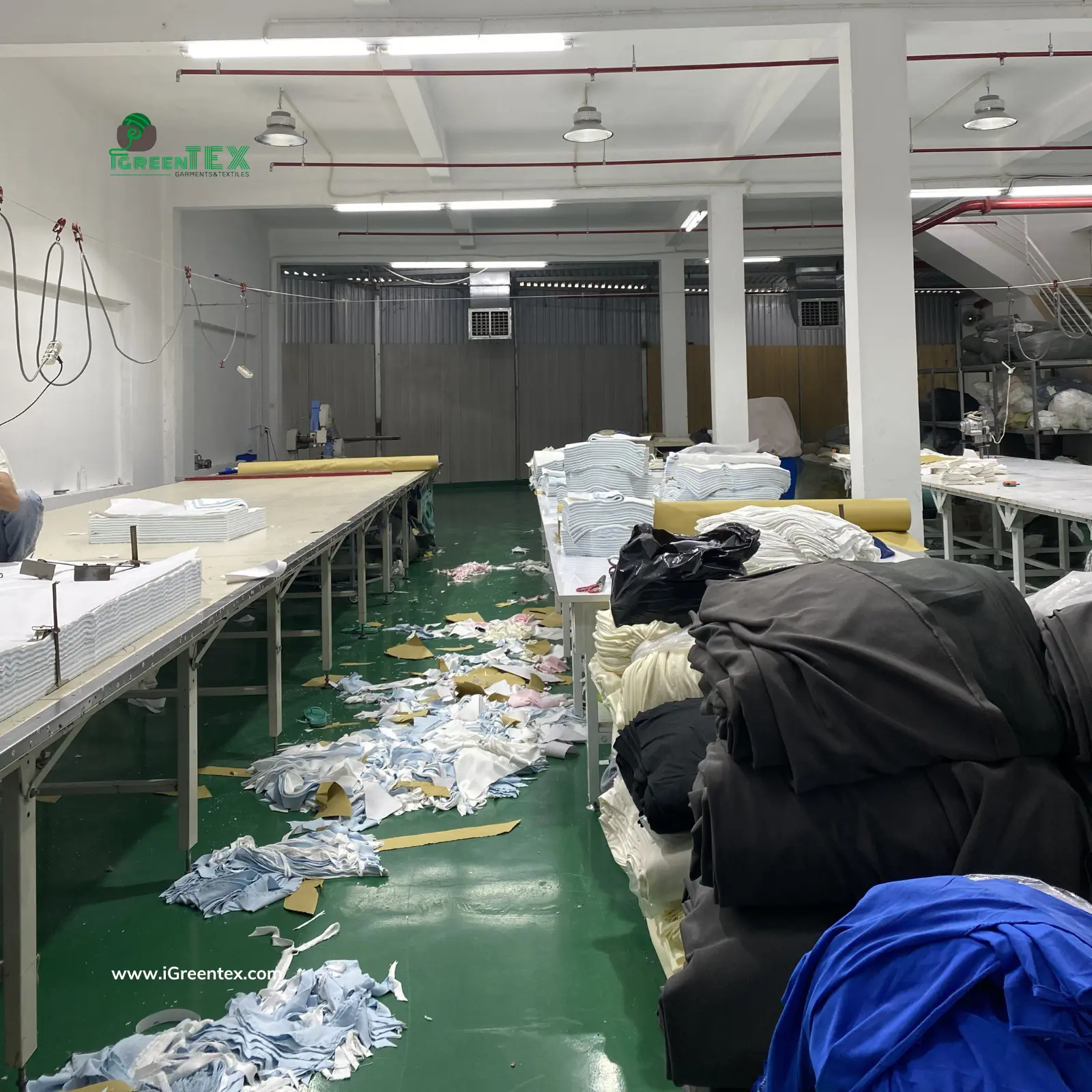 An authentic documentary-style photograph capturing a Vietnamese garment worker actively cutting blue denim fabric at one of the main cutting tables within the bustling iGreen Tex factory in Vietnam. The image highlights precise manual fabric cutting for bulk apparel orders, showcasing internal production capacity and efficient manufacturing workflow.