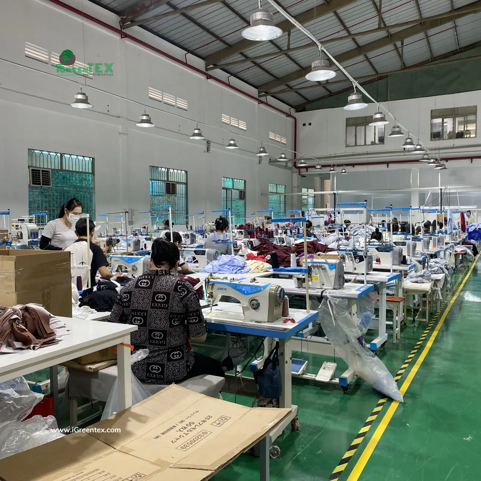 A dynamic, photorealistic view of multiple sewing lines operating at full capacity within iGreen Tex Vietnam's apparel factory, demonstrating our real production capability. Large stacks of semi-finished garments are visible, signifying high-volume export orders.