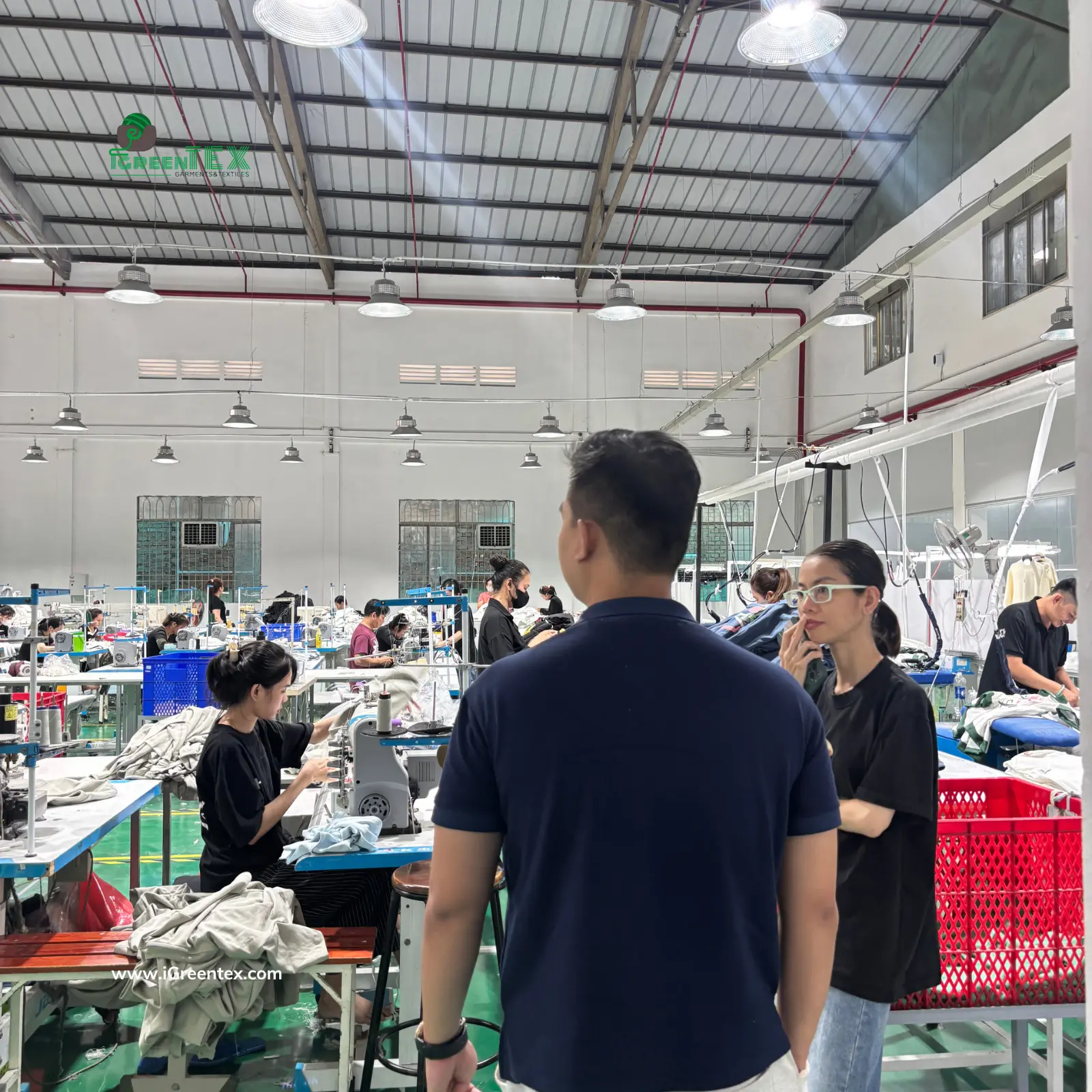 A professional supply chain manager leads a Western apparel buyer delegation through the final quality control and packing area at iGreen Tex to verify garment factory vietnam compliance. Both buyers are carefully examining finished products and observing meticulous inspection standards, providing real visual proof to help them verify garment factory vietnam partners.