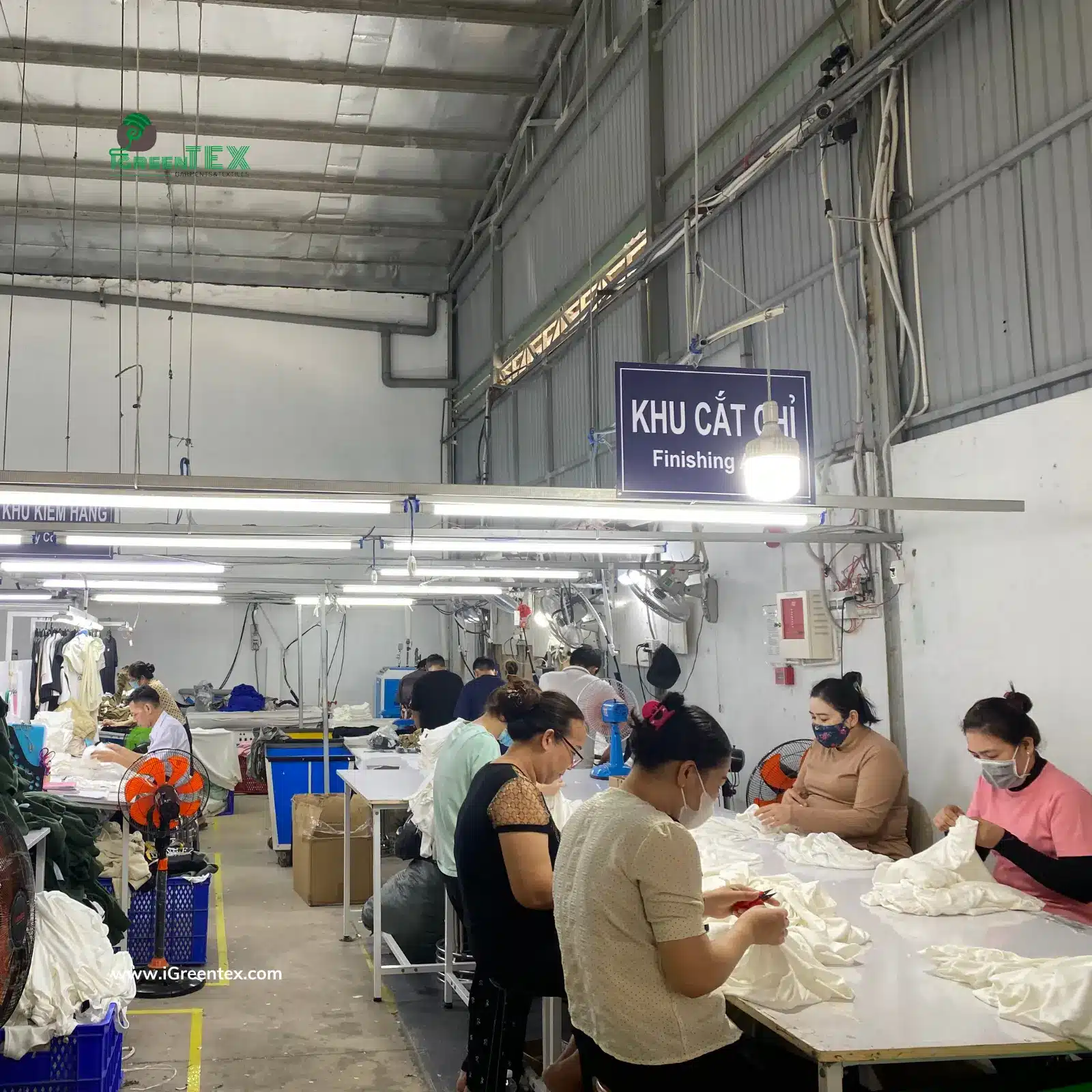 An authentic photograph from the finishing department at iGreen Tex in Vietnam, showing skilled workers carefully cutting excess threads from finished garments. This meticulous manual process highlights a key advantage when comparing Vietnam vs China manufacturing cost: achieving high-quality finishing at competitive labor rates.